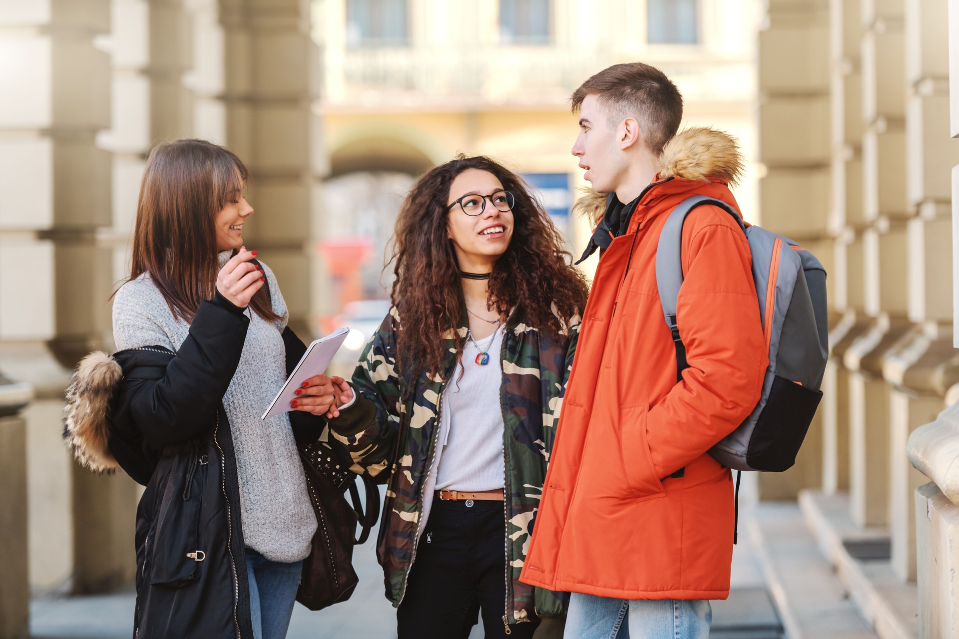 Three smiling happy multicultural friends standing in front of school and chatting.